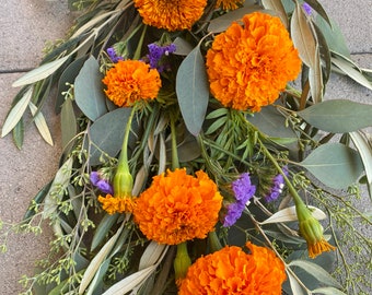 May include: A floral arrangement featuring vibrant orange marigold blooms, interspersed with delicate purple statice and green foliage. The arrangement includes eucalyptus leaves and olive branches, creating a natural and textured aesthetic.