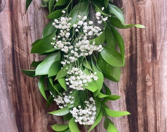 May include: A garland of white baby's breath flowers and green leaves hanging against a wooden background.