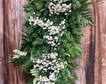 May include: A garland of white baby's breath flowers and green ferns hanging on a wooden wall.