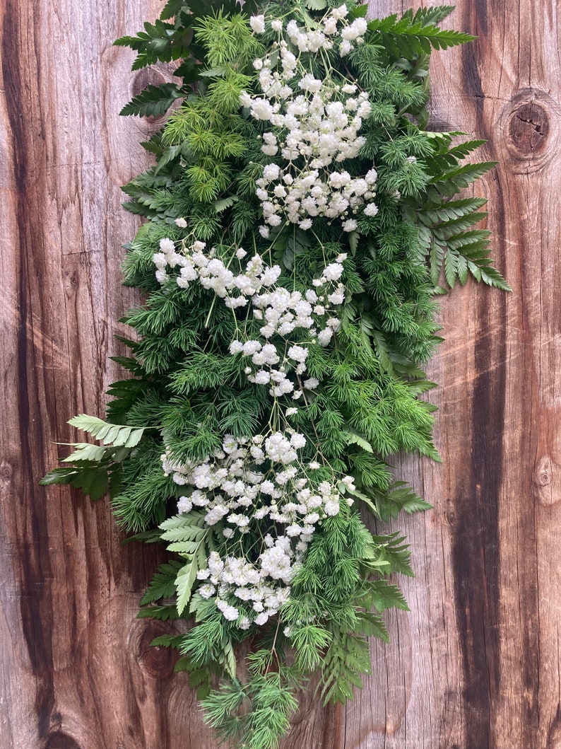 以下が含まれることがあります： 木製の壁に白いカスミソウの花と緑のシダのガーランドが飾られています