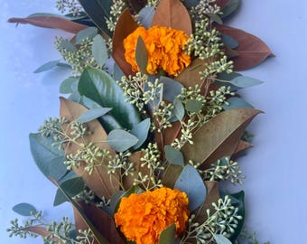 May include: A floral arrangement featuring three vibrant orange marigold blooms. The arrangement includes large, dark green leaves, brown leaves, and small, light green buds. The flowers and foliage are arranged in a linear fashion.