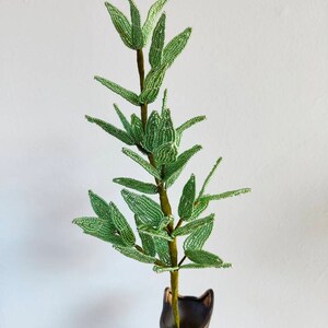 May include: A green beaded plant with leaves in a black vase. The plant is made of small green beads and is a realistic representation of a leafy plant.