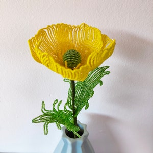 K&ouml;nnte beinhalten: Eine gelbe Perlenblume mit gr&uuml;nen Perlenbl&auml;ttern in einer hellblauen Vase.