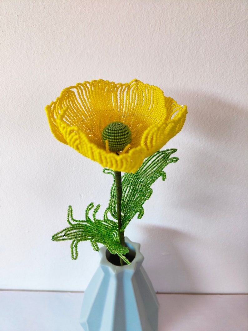 K&ouml;nnte beinhalten: Eine gelbe Perlenblume mit gr&uuml;nen Perlenbl&auml;ttern in einer hellblauen Vase.