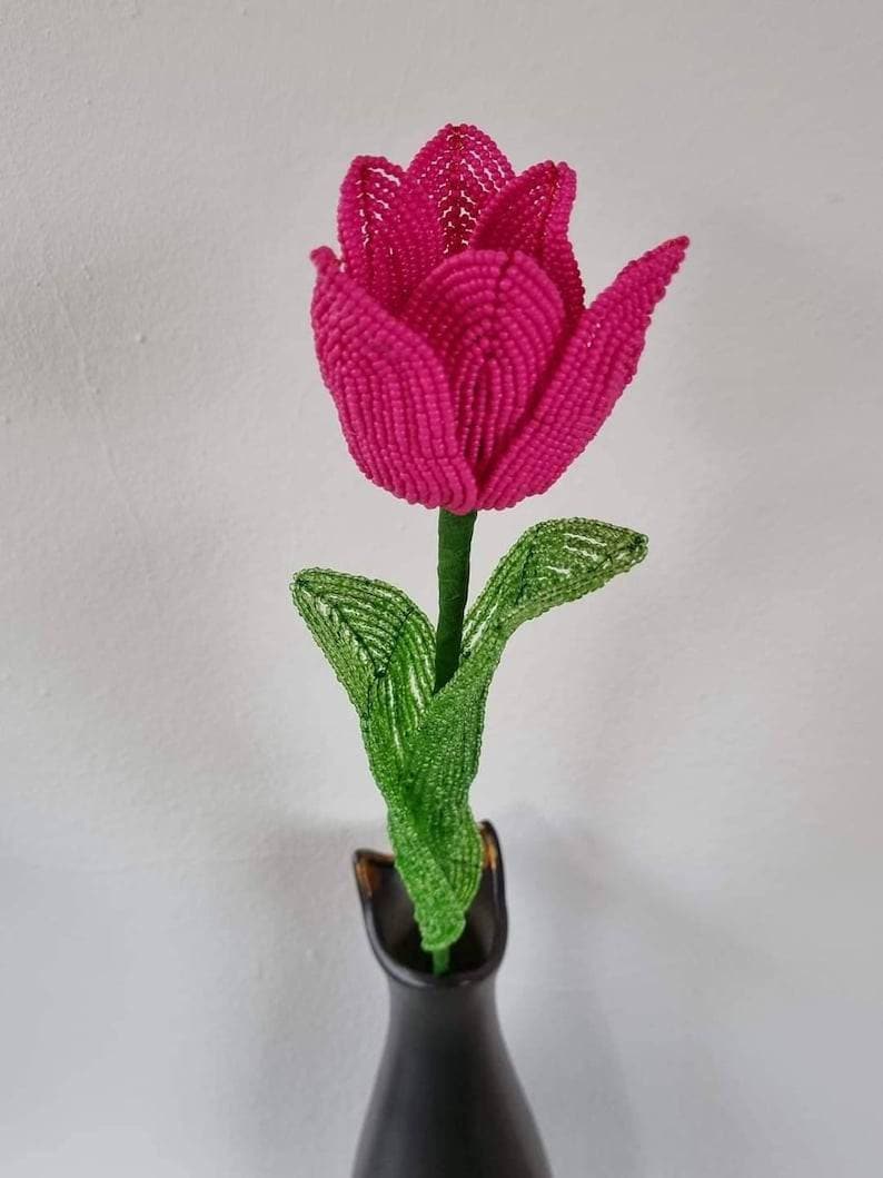 May include: A pink beaded tulip flower with green beaded leaves in a black vase.