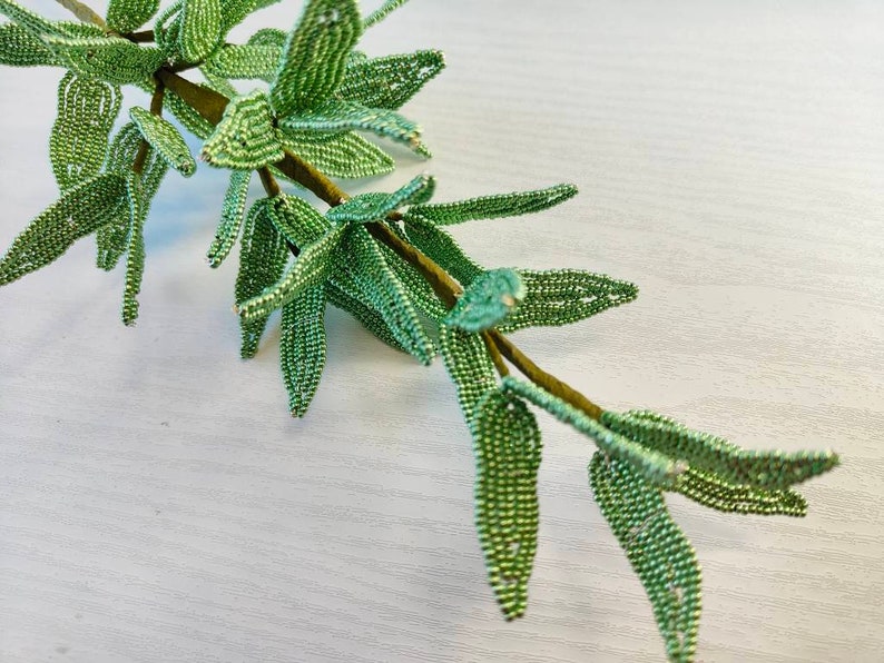 May include: A green beaded branch with leaves, crafted with small, round beads. The branch is on a white background.