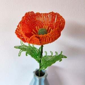 K&ouml;nnte beinhalten: Eine Perlen-Mohnblume in Orange- und Gelbt&ouml;nen mit gr&uuml;nen Bl&auml;ttern, arrangiert in einer hellblauen Vase.