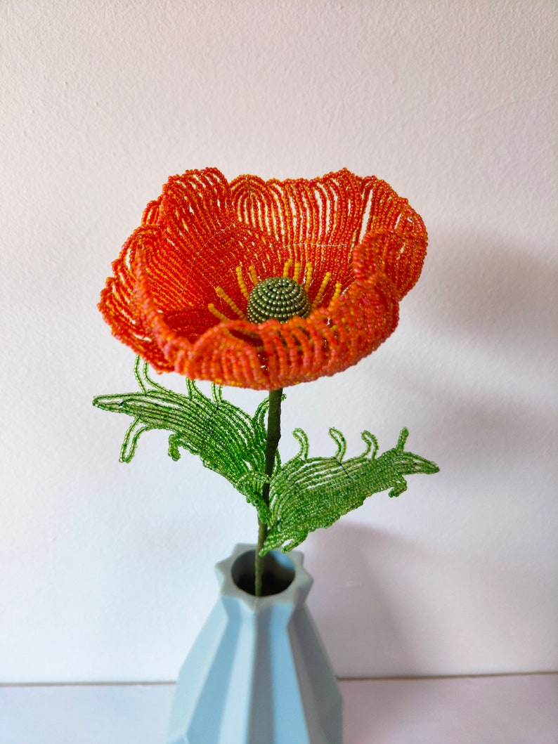 K&ouml;nnte beinhalten: Eine Perlen-Mohnblume in Orange- und Gelbt&ouml;nen mit gr&uuml;nen Bl&auml;ttern, arrangiert in einer hellblauen Vase.