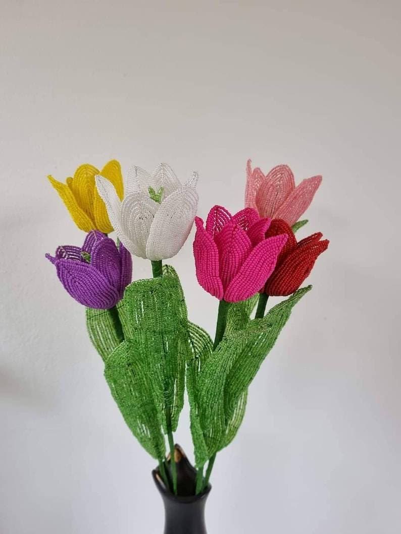 May include: A bouquet of five beaded tulips in a vase. The tulips are white, yellow, purple, pink, and red. The leaves are green.