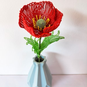 K&ouml;nnte beinhalten: Eine rote Perlen-Mohnblume mit einem gr&uuml;nen Perlenstiel und Bl&auml;ttern in einer hellblauen Vase.