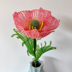 K&ouml;nnte beinhalten: Eine rosa Perlenblume mit gr&uuml;nen Bl&auml;ttern in einer hellblauen Vase. Die Blume besteht aus kleinen Perlen und hat eine gelbe Mitte.