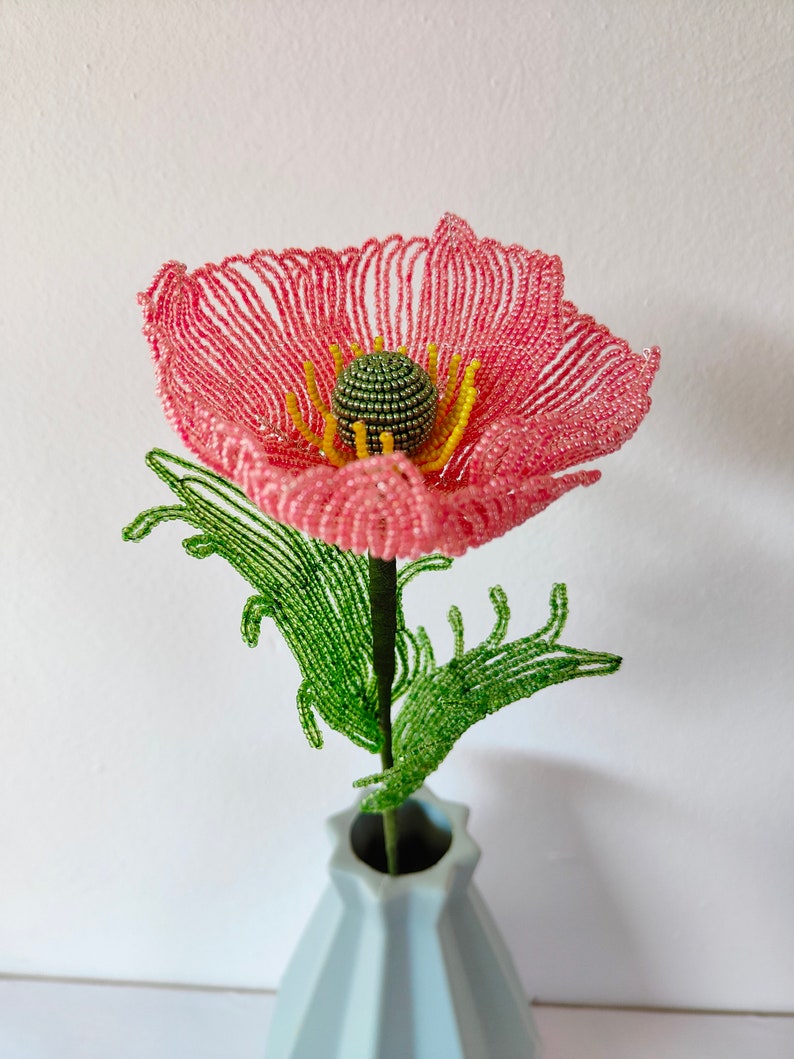 K&ouml;nnte beinhalten: Eine rosa Perlenblume mit gr&uuml;nen Bl&auml;ttern in einer hellblauen Vase. Die Blume besteht aus kleinen Perlen und hat eine gelbe Mitte.