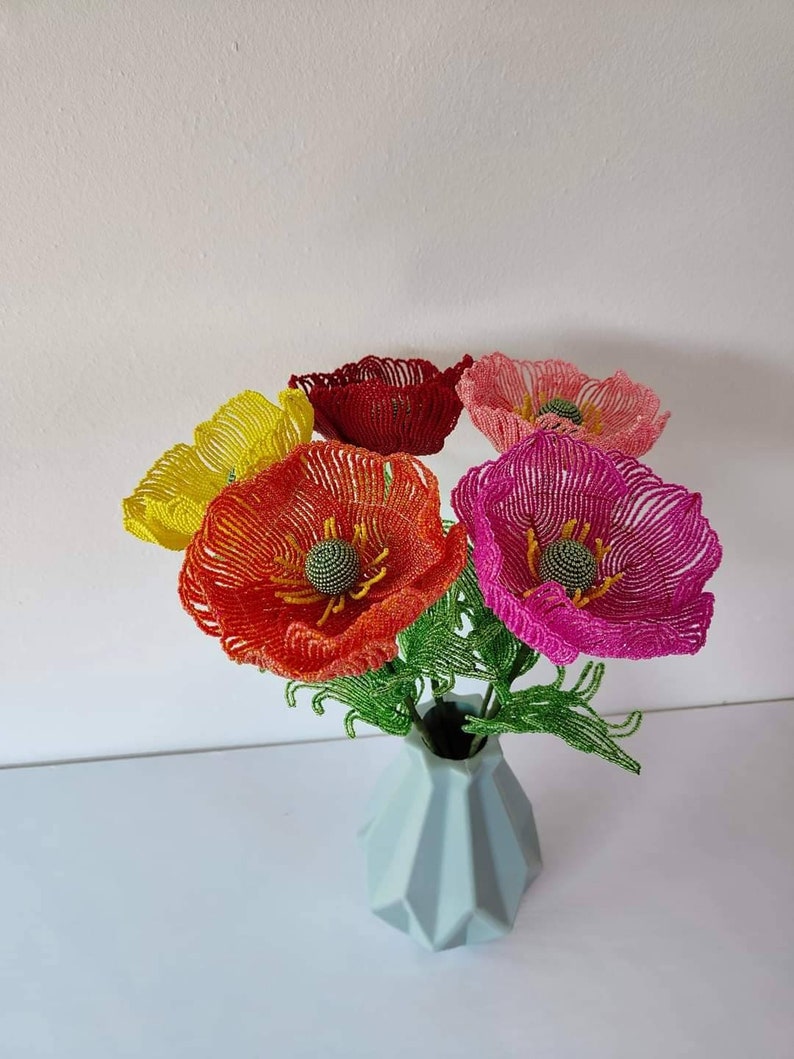 K&ouml;nnte beinhalten: Ein Strau&szlig; aus f&uuml;nf Perlen-Mohnblumen in einer hellblauen Vase. Die Blumen sind rot, orange, gelb, rosa und dunkelrosa. Die Blumen sind aus kleinen Perlen gefertigt und haben ein realistisches Aussehen.