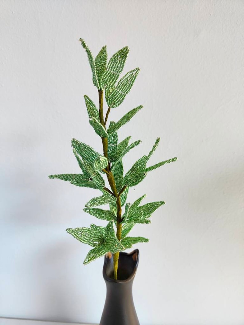 May include: A green beaded plant with a brown stem in a black vase. The plant is made of small green beads and has a realistic appearance.