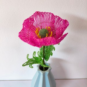 K&ouml;nnte beinhalten: Eine rosa Perlenblume mit gr&uuml;nen Bl&auml;ttern in einer hellblauen Vase. Die Blume ist aus kleinen Perlen gefertigt und hat einen zarten, femininen Look.