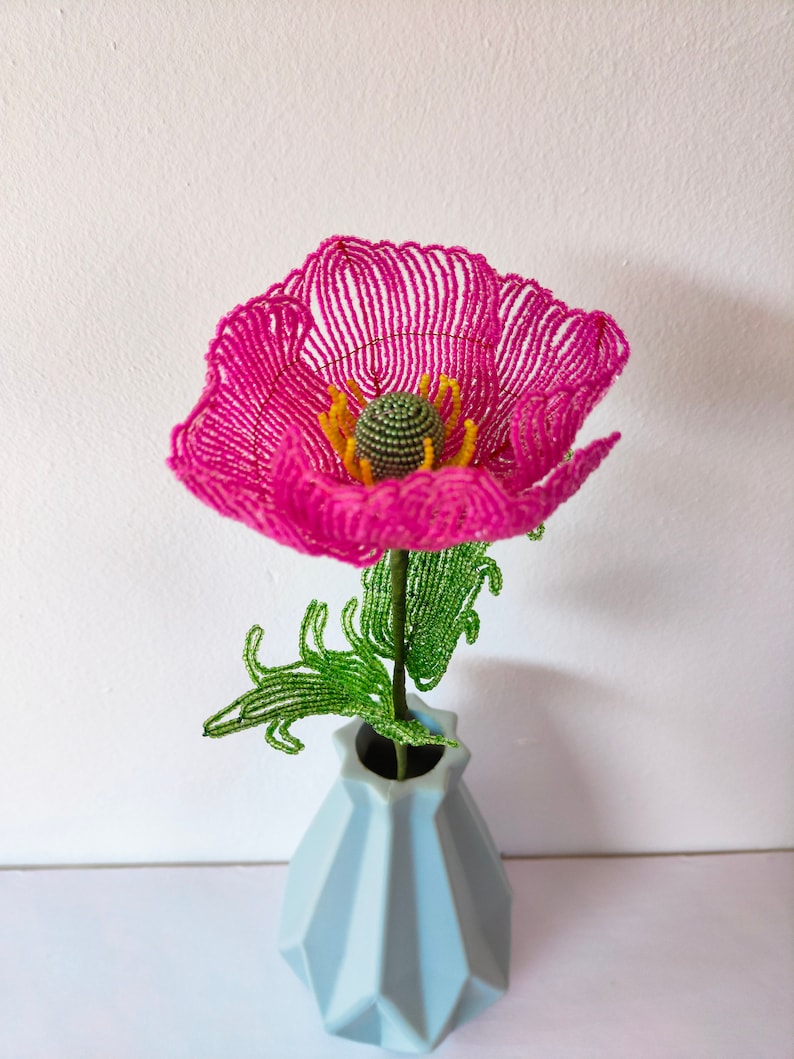 K&ouml;nnte beinhalten: Eine rosa Perlenblume mit gr&uuml;nen Bl&auml;ttern in einer hellblauen Vase. Die Blume ist aus kleinen Perlen gefertigt und hat einen zarten, femininen Look.