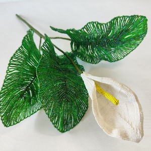 May include: A white beaded flower with a yellow center and green beaded leaves. The flower is made of small white beads and the leaves are made of small green beads.