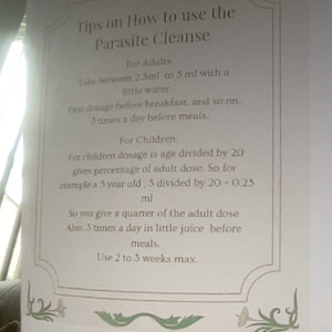 May include: A white sheet of paper with black text outlining instructions on how to use a parasite cleanse. The text includes dosage information for adults and children, as well as the frequency and duration of use. The paper has a decorative border with green floral designs.