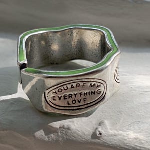 Peut inclure: Bague argentée avec un bord ondulé et une bordure en émail vert. La bague porte l'inscription gravée "YOU ARE MY EVERYTHING LOVE" sur une plaque ovale. La bague a un aspect vintage et usé.