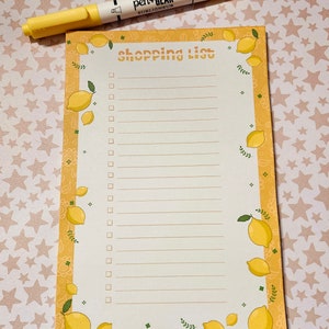 May include: A lemon-themed shopping list notepad with a yellow border and lemon illustrations. The notepad has a white background with lines for writing and the words "shopping list" at the top. A yellow highlighter pen is placed on top.