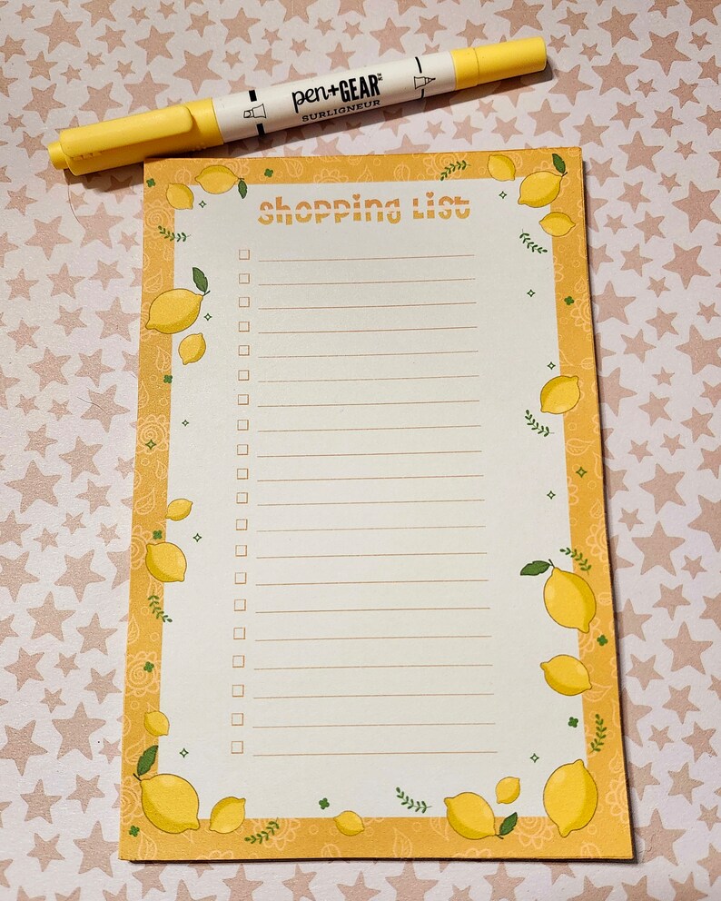 May include: A lemon-themed shopping list notepad with a yellow border and lemon illustrations. The notepad has a white background with lines for writing and the words "shopping list" at the top. A yellow highlighter pen is placed on top.