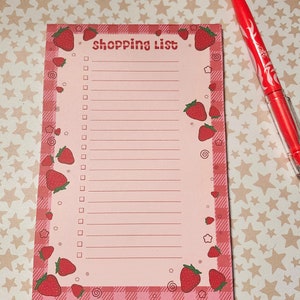 May include: A pink shopping list notepad with a strawberry theme. The notepad has a gingham border and is decorated with red strawberries. The words "shopping list" are printed at the top. A red pen is next to the notepad.