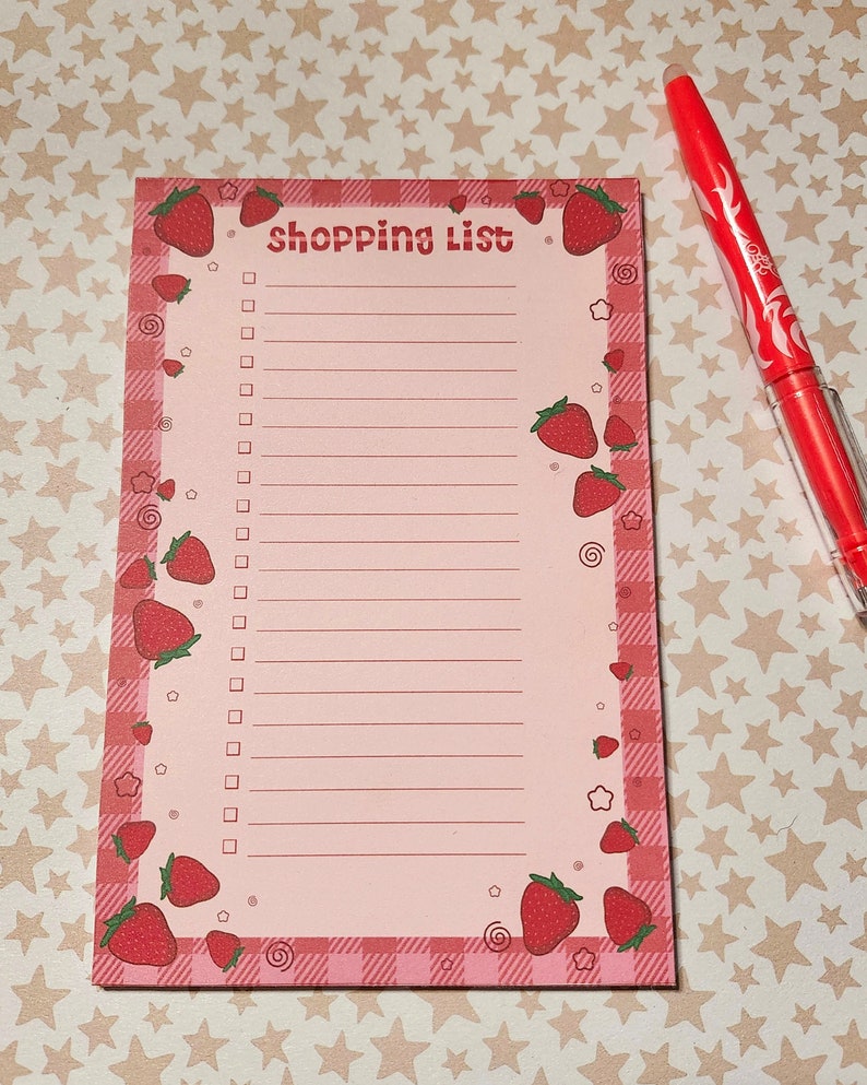 May include: A pink shopping list notepad with a strawberry theme. The notepad has a gingham border and is decorated with red strawberries. The words "shopping list" are printed at the top. A red pen is next to the notepad.