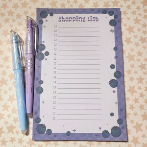 May include: A shopping list notepad with a blueberry theme, featuring a purple checkered border and blueberry illustrations. Two pens, one light blue and one lavender, are positioned to the left of the notepad.