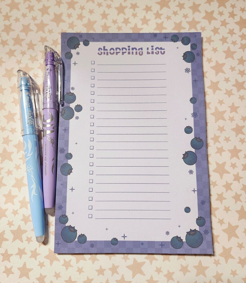 May include: A shopping list notepad with a blueberry theme, featuring a purple checkered border and blueberry illustrations. Two pens, one light blue and one lavender, are positioned to the left of the notepad.