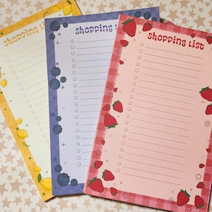 May include: Three colorful shopping list notepads. One is yellow with lemons, another is purple with blueberries, and the third is pink with strawberries. Each notepad has a decorative border and the words "shopping list" at the top.