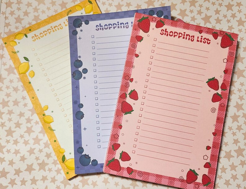 May include: Three colorful shopping list notepads. One is yellow with lemons, another is purple with blueberries, and the third is pink with strawberries. Each notepad has a decorative border and the words "shopping list" at the top.