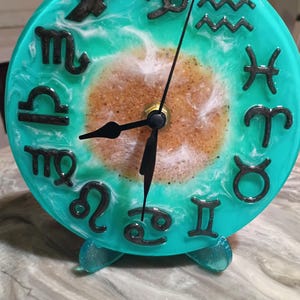May include: A round clock featuring a turquoise and white marbled resin face with a central sandy-textured circle. Twelve black raised zodiac symbols are arranged around the perimeter, with black hour, minute, and second hands at the center.