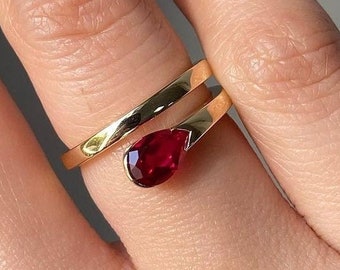 May include: A gold ring with a red gemstone in the shape of a teardrop. The ring is wrapped around the finger.