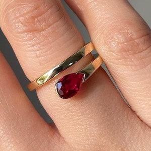 May include: A gold ring with a red gemstone in the shape of a teardrop. The ring is wrapped around the finger.