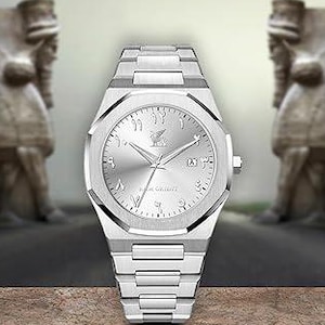 May include: A silver-toned wristwatch with a metallic band and a round face. The watch features Arabic numerals and a logo at the 12 o'clock position. The background includes stone sculptures and a blurred road.