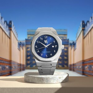May include: A silver-toned wristwatch with a blue dial and Arabic numerals. The watch features a date display and a logo of a winged creature. The watch is displayed on a small, round, concrete pedestal.