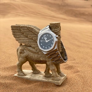 May include: A silver wristwatch with a black face and Arabic numerals rests on a stone winged bull statue. The watch has a metallic band and is set against a desert backdrop.