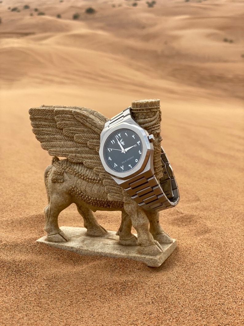 May include: A silver wristwatch with a black face and Arabic numerals rests on a stone winged bull statue. The watch has a metallic band and is set against a desert backdrop.