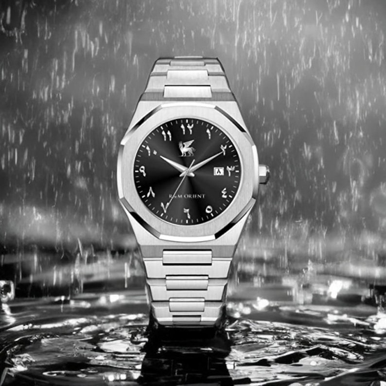 K&ouml;nnte beinhalten: Silberne Armbanduhr mit schwarzem Zifferblatt und arabischen Ziffern. Die Uhr hat ein Edelstahlarmband und ein rundes Zifferblatt mit dem Markennamen "R & M Orient". Der Hintergrund zeigt verschwommene Wassertropfen.