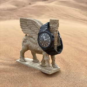 May include: A black wristwatch with Arabic numerals and a matching black band is displayed on a stone sculpture of a winged bull. The watch face is dark with light-colored hands and text. The background is a desert landscape with sand dunes.