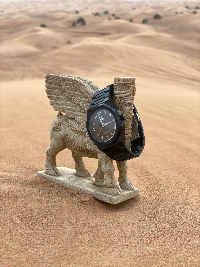 K&ouml;nnte beinhalten: Eine schwarze Armbanduhr mit arabischen Ziffern und einem passenden schwarzen Armband wird auf einer Steinskulptur eines gefl&uuml;gelten Stiers pr&auml;sentiert. Das Zifferblatt der Uhr ist dunkel mit hellen Zeigern und Text. Der Hintergrund ist eine W&uuml;stenlandschaft.