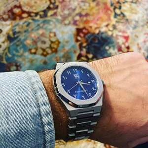 May include: A stainless steel wristwatch with a blue face and Arabic numerals. The watch has a geometric design and a metal band. The text "R&M ORIENT" is visible on the watch face.