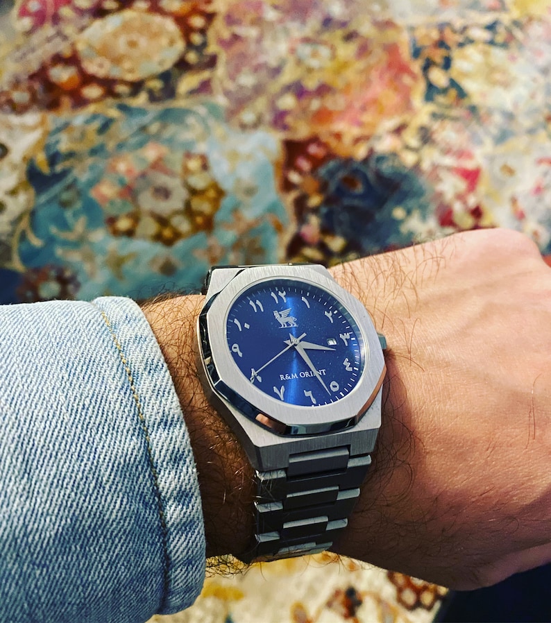 May include: A stainless steel wristwatch with a blue face and Arabic numerals. The watch has a geometric design and a metal band. The text "R&M ORIENT" is visible on the watch face.
