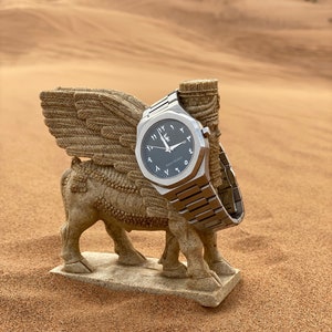 K&ouml;nnte beinhalten: Eine silberne Armbanduhr mit dunkelgrauem Zifferblatt und arabischen Ziffern liegt auf einer Steinskulptur eines gefl&uuml;gelten Stiers. Die Uhr hat ein Metallarmband, vor W&uuml;stenhintergrund.