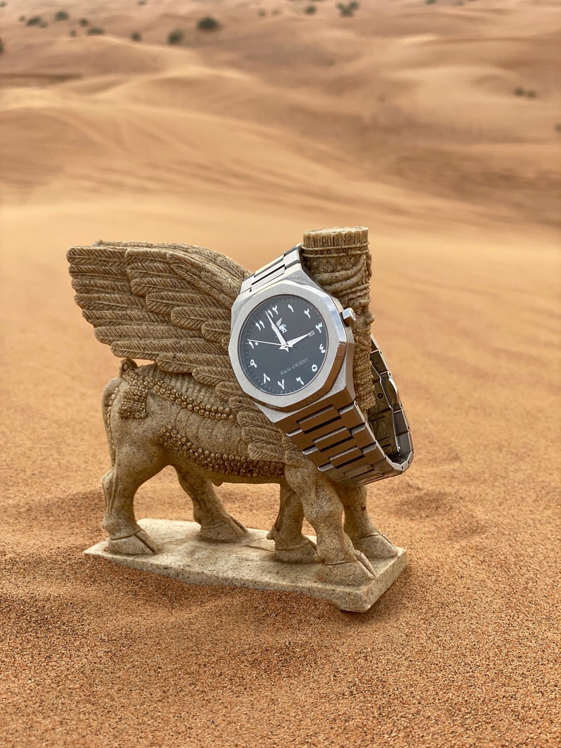 K&ouml;nnte beinhalten: Eine silberne Armbanduhr mit dunkelgrauem Zifferblatt und arabischen Ziffern liegt auf einer Steinskulptur eines gefl&uuml;gelten Stiers. Die Uhr hat ein Metallarmband, vor W&uuml;stenhintergrund.