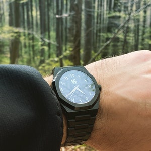 K&ouml;nnte beinhalten: Eine schwarze Armbanduhr mit geometrischem Design. Das Zifferblatt hat arabische Ziffern und ein Logo. Die Uhr hat ein schwarzes Armband. Der Hintergrund ist ein Wald.