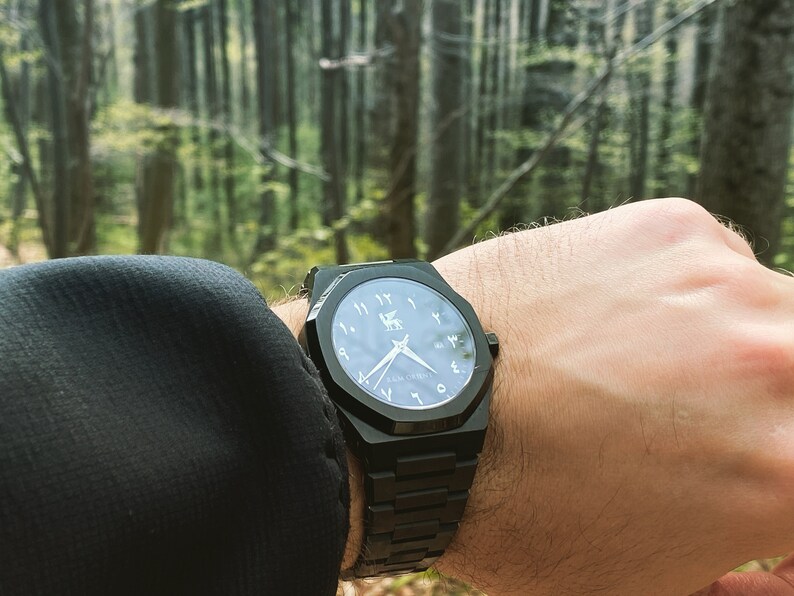 K&ouml;nnte beinhalten: Eine schwarze Armbanduhr mit geometrischem Design. Das Zifferblatt hat arabische Ziffern und ein Logo. Die Uhr hat ein schwarzes Armband. Der Hintergrund ist ein Wald.