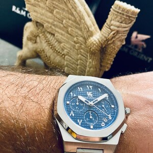 K&ouml;nnte beinhalten: Eine Edelstahl-Armbanduhr mit blauem Zifferblatt und arabischen Ziffern. Die Uhr hat ein Chronographen-Design mit mehreren Zifferbl&auml;ttern und ein silbernes Armband. Die Uhr ist mit "R&M Orient" gekennzeichnet. Im Hintergrund eine beige Skulptur einer gefl&uuml;gelten Figur.