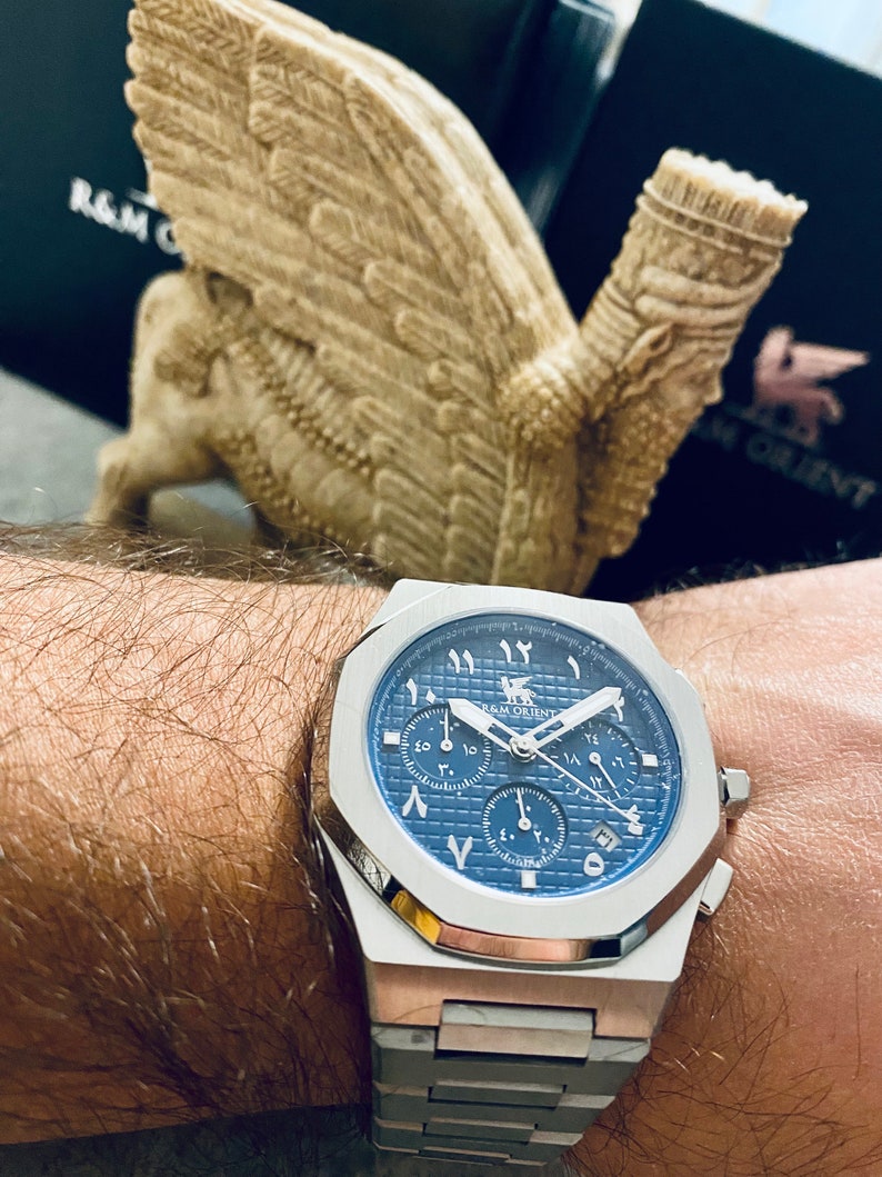 K&ouml;nnte beinhalten: Eine Edelstahl-Armbanduhr mit blauem Zifferblatt und arabischen Ziffern. Die Uhr hat ein Chronographen-Design mit mehreren Zifferbl&auml;ttern und ein silbernes Armband. Die Uhr ist mit "R&M Orient" gekennzeichnet. Im Hintergrund eine beige Skulptur einer gefl&uuml;gelten Figur.