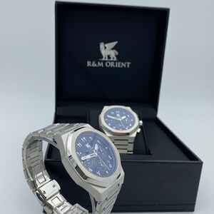 K&ouml;nnte beinhalten: Eine Edelstahl-Armbanduhr mit blauem Zifferblatt und arabischen Ziffern wird neben ihrer Box pr&auml;sentiert. Die Uhr hat ein passendes Edelstahlarmband und wird auf einem transparenten St&auml;nder pr&auml;sentiert. Die Box ist schwarz mit dem Markennamen "R&M ORIENT".